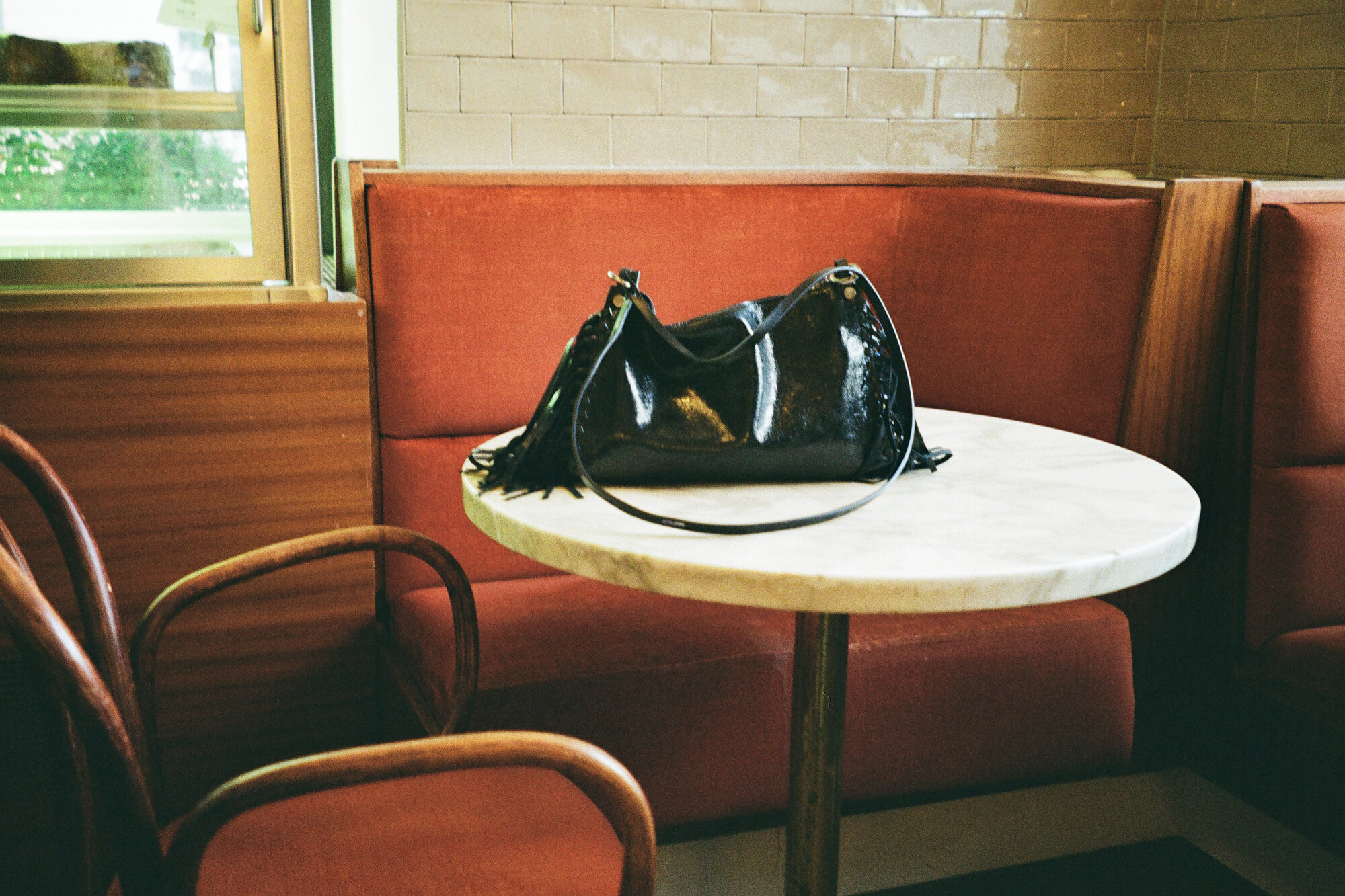 Eine schwarze Handtasche mit Fransen, die auf einem runden Café-Tisch mit Marmorplatte vor einer roten Sitzecke steht.