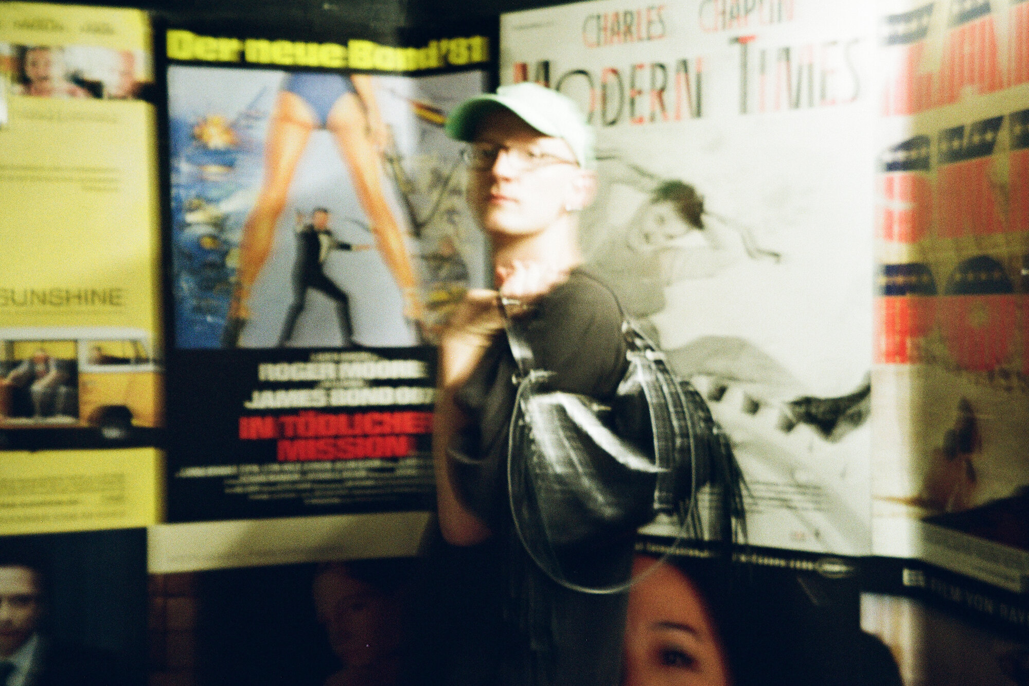 Eine Person mit einer hellgrünen Mütze und einer Brille steht vor mehreren alten Filmplakaten.