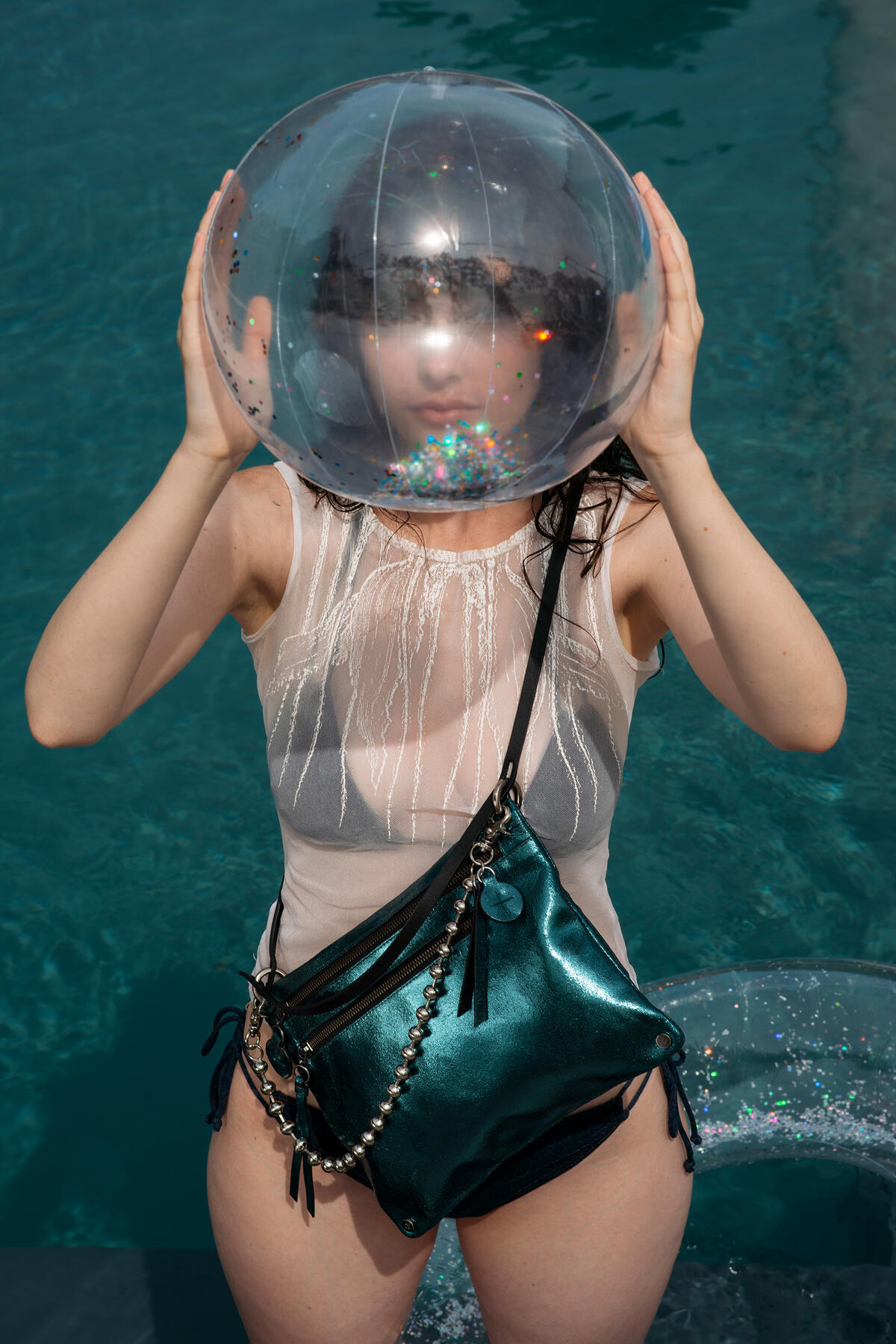 Ein Mädchen mit Anfang zwanzig steht in einem Pool mit einer INA KENT-Tasche MOONLIT ed.2 aus blaugrünem Metallic-Leder und einer Schlüsselkette umgehängt. Sie hält sich einen durchsichtigen Wasserball vors Gesicht.