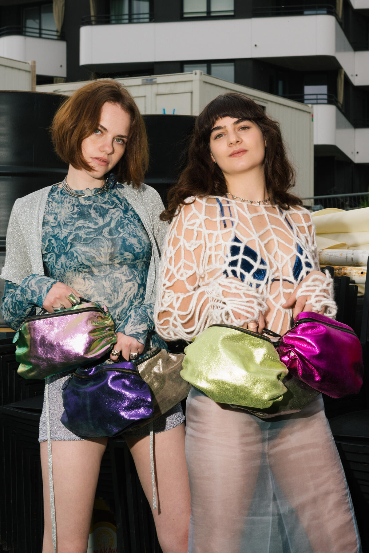 Two women standing in front of a building, holding shiny, colorful INA KENT drawstring bags. They are wearing stylish outfits and looking directly at the camera.