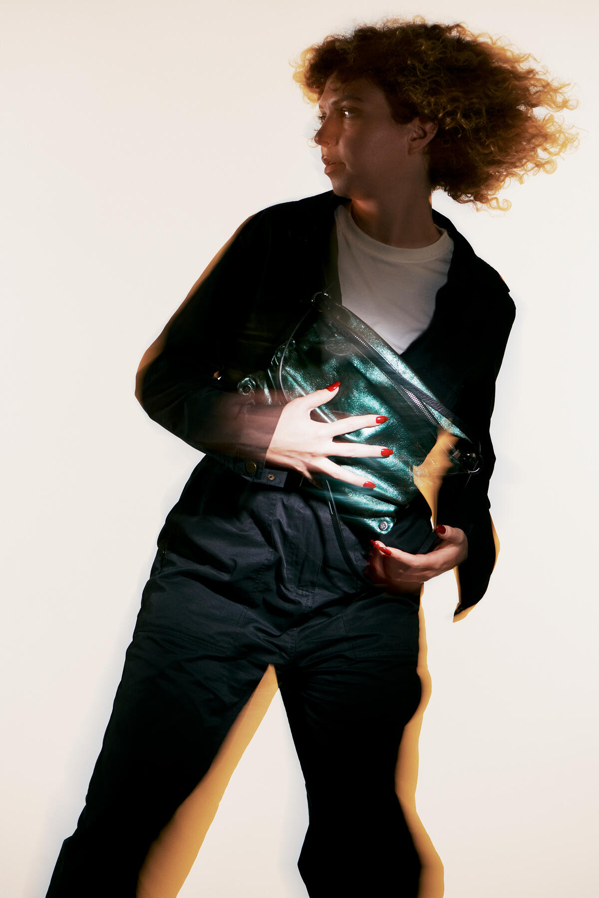 A person with curly hair holds a shiny green INA KENT bag against a light background. They wear a dark jacket, pants, and a white shirt. The image has a blurred, artistic effect.