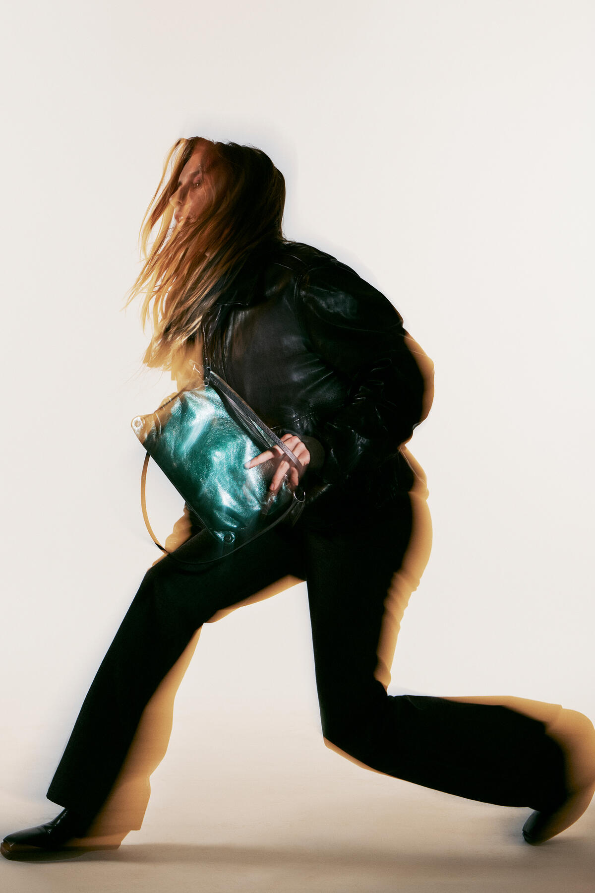 A person in motion, wearing a black jacket and pants, clutches a stylish INA KENT green handbag. The image captures the essence of dynamic movement with its blur effect.