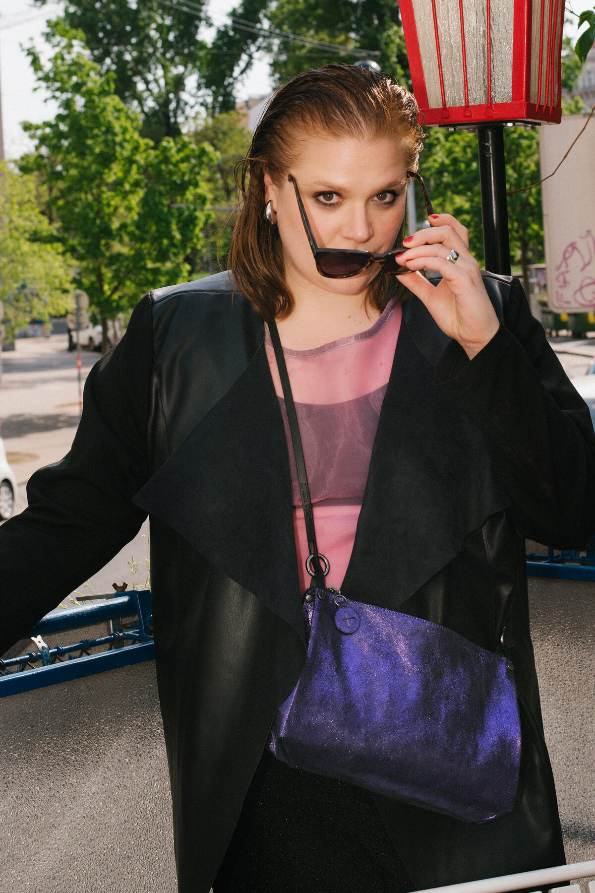 A person with sunglasses and a phone, wearing a black jacket, purple top, and an INA KENT purple purse, stands outdoors with greenery in the background.