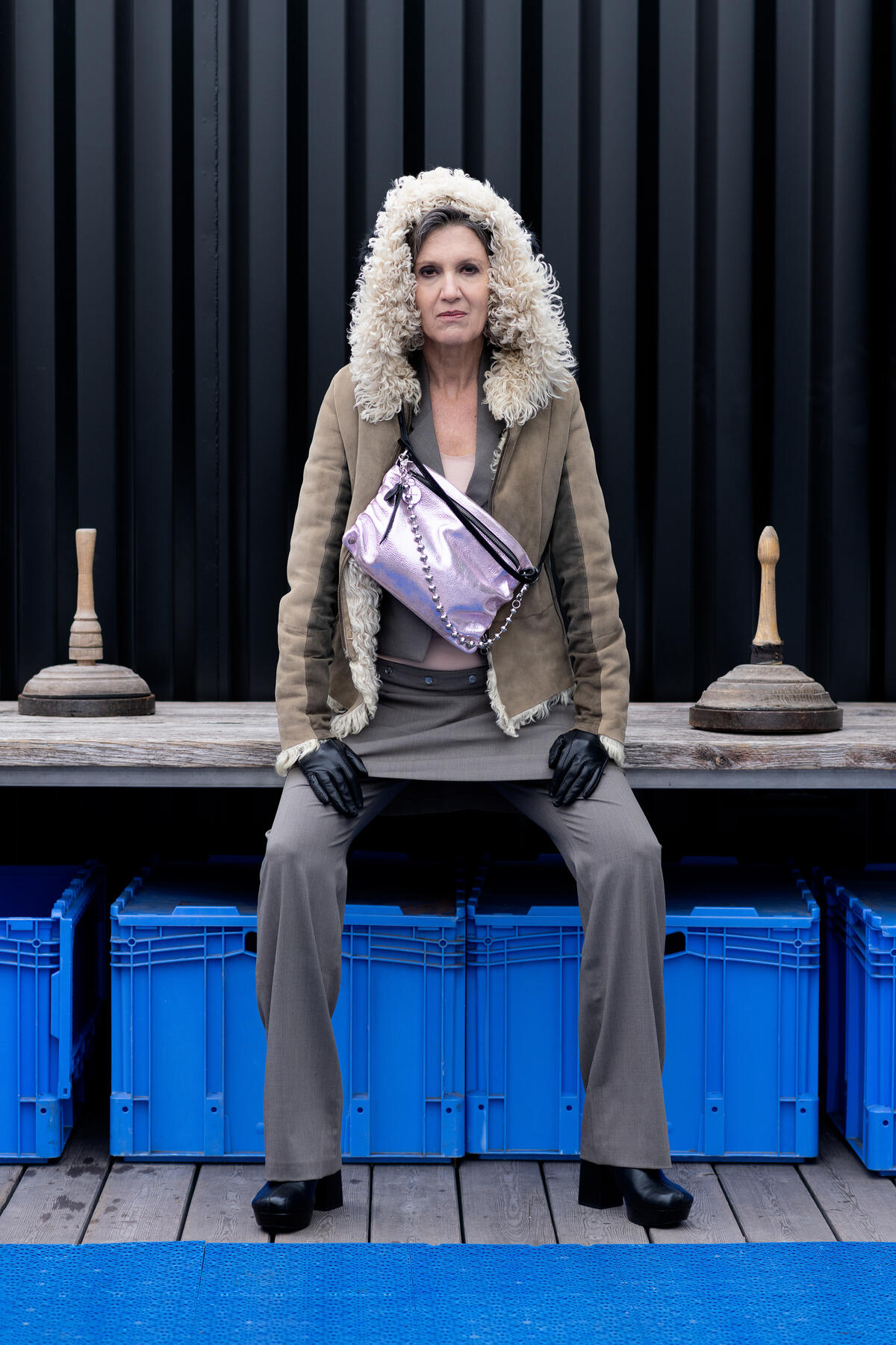  Die Frau trägt eine Metallic-Lavender-Rose-Tasche von INA KENT als Crossbody-Bag über einer olivfarbenen Lammfelljacke. Sie sitzt auf einer Holzbank vor einer schwarzen Containerwand mit blauen Kisten darunter. Die Tasche ist mit einer Kettenriemen-Option versehen, die ihr einen eleganten Touch verleiht.