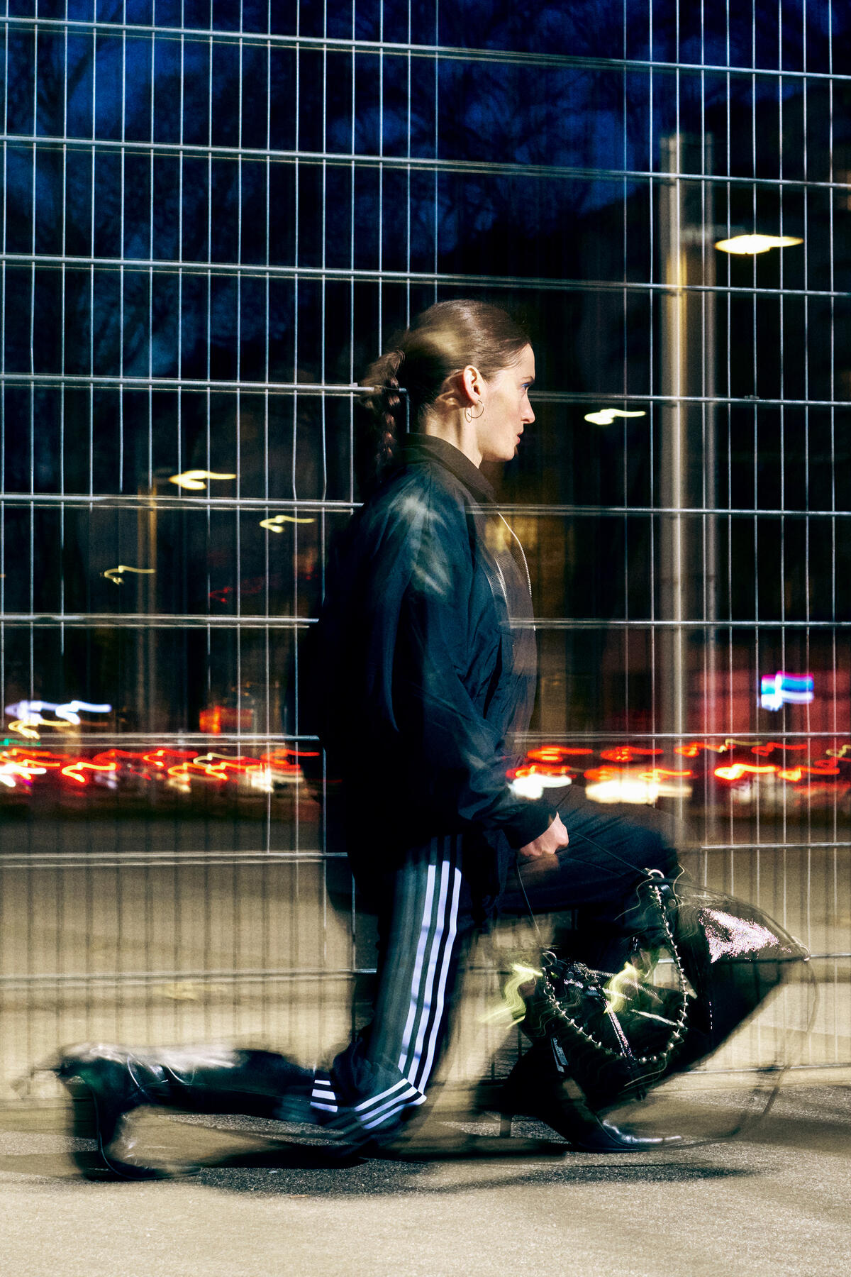 A person in athletic wear and boots kneels beside a metal fence at night, holding a shiny INA KENT bag, with blurred lights and motion creating a dynamic effect.