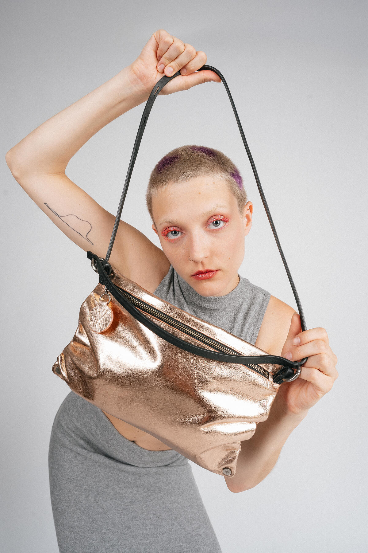 Person with short hair and pink eye makeup holds up a metallic gold INA KENT handbag by its strap, wearing a sleeveless gray outfit against a plain background.