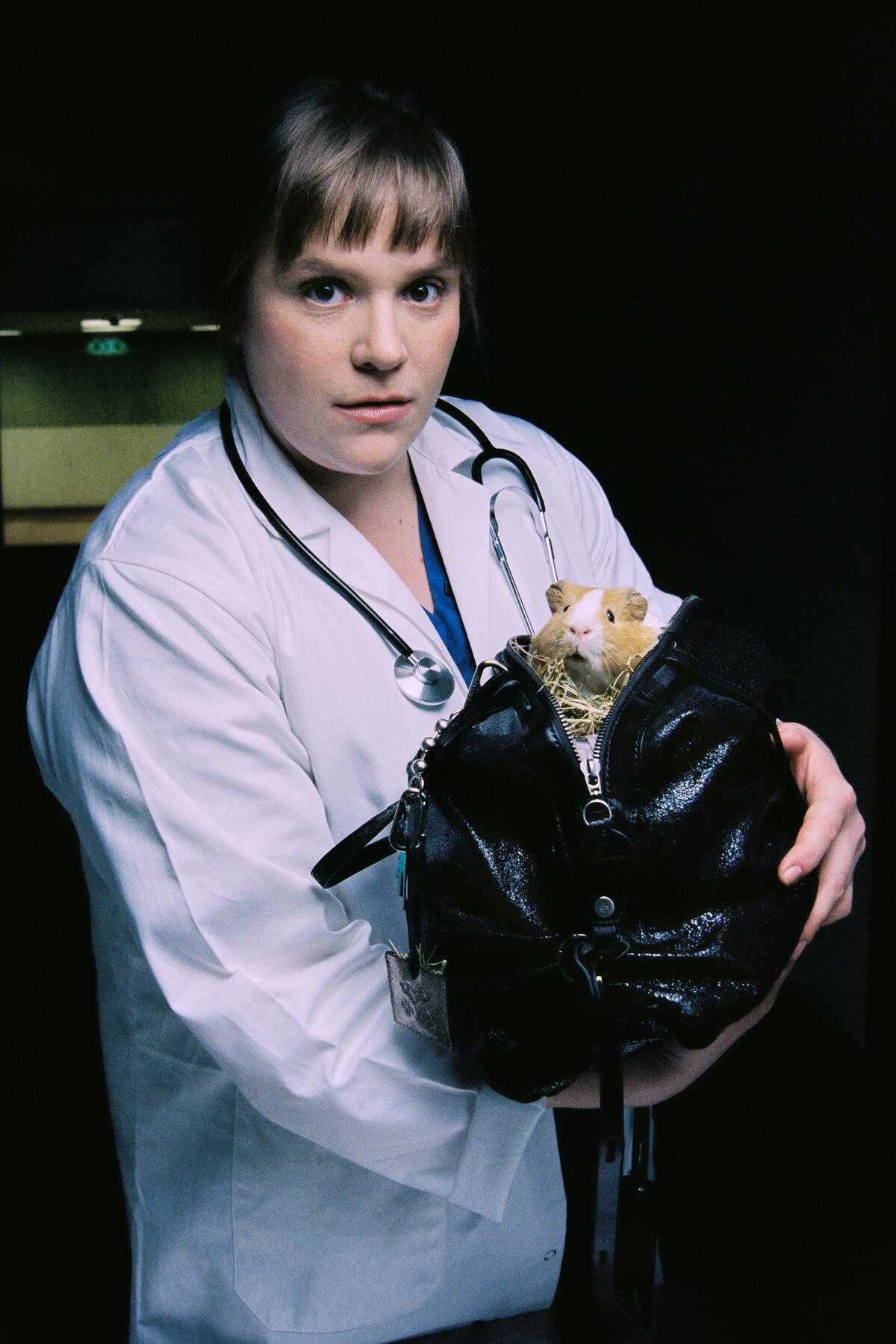 A person in a white lab coat with a stethoscope holds a sleek INA KENT black bag, partially unzipped to reveal a guinea pig inside.