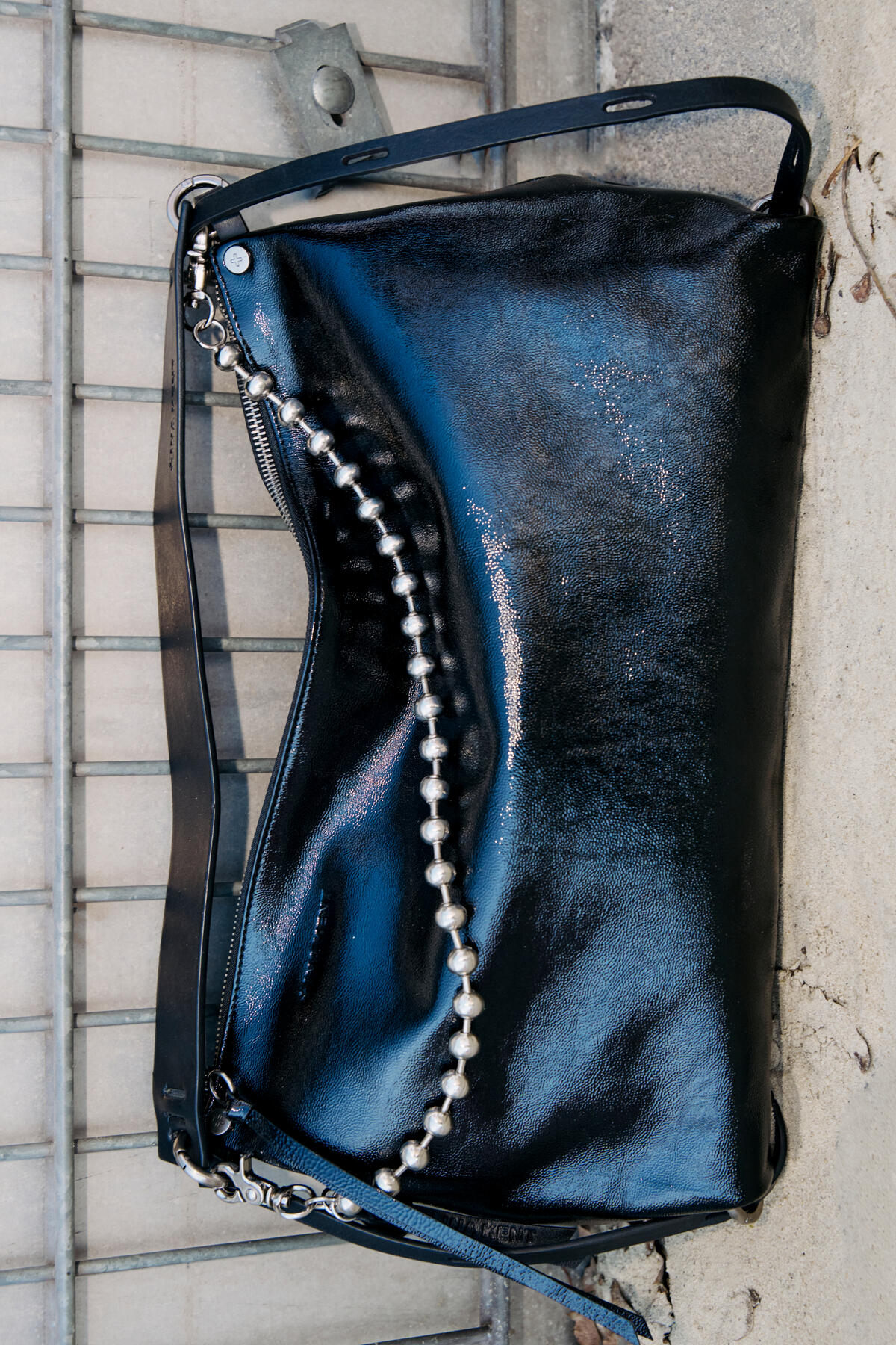 An INA KENT black patent leather handbag with a silver chain and pearl detail rests on a concrete surface next to a metal fence.
