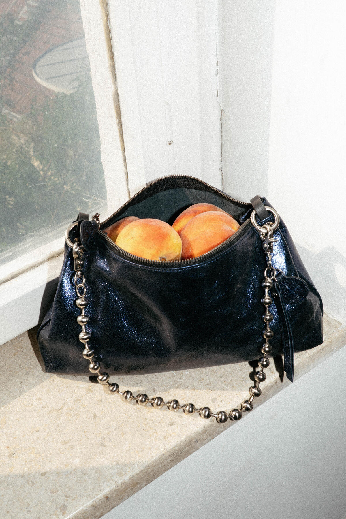 Eine glänzende schwarze Handtasche von INA KENT mit einem Kettenriemen liegt auf einer Fensterbank, der Reißverschluss ist geöffnet und gibt den Blick auf mehrere Pfirsiche frei.