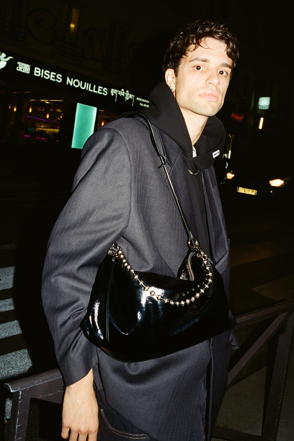 Ein Mann in schwarzem Kapuzenpulli und übergroßem Blazer steht nachts vor der Tür und trägt eine glänzende schwarze Handtasche von INA KENT mit einem Kettenriemen über der Schulter.
