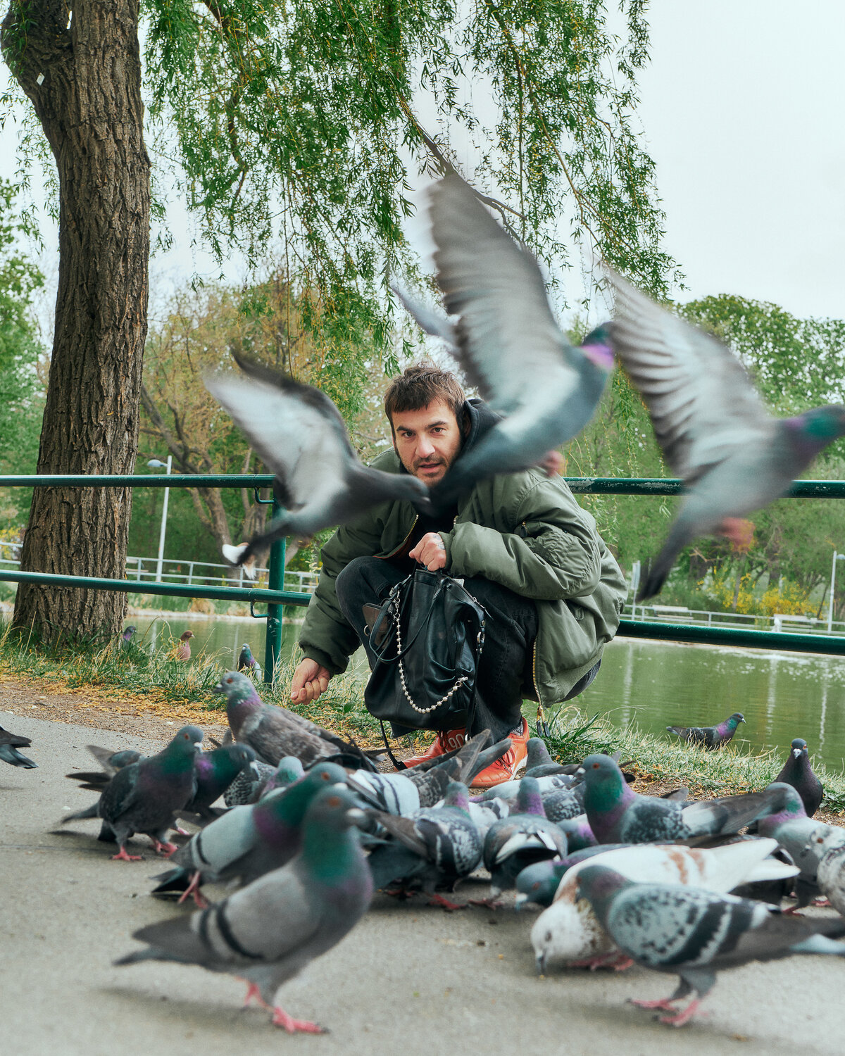 A man crouches by a pond feeding pigeons, with one pigeon in mid-flight in front of him and several more gathered on the ground, his INA KENT bag resting at his side.