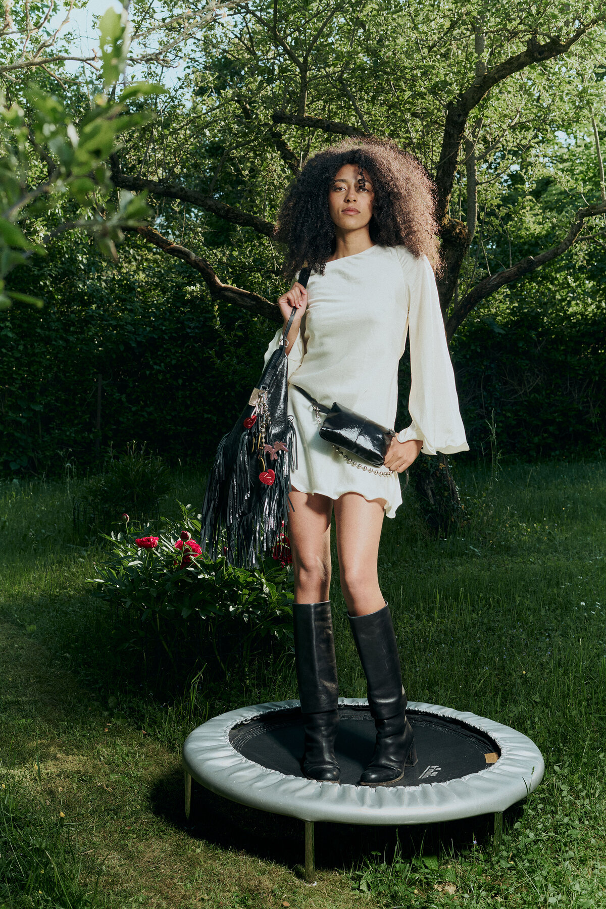 A woman in a white dress and black boots stands on a trampoline in a garden, holding an INA KENT black bag in one hand and a clutch in the other.