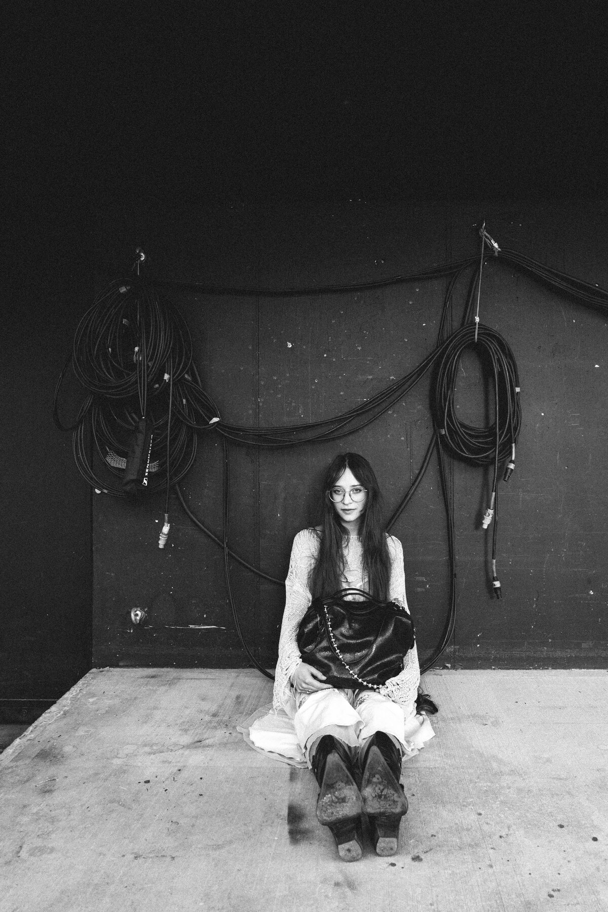 A woman in glasses and boots sits on the ground against a dark wall with coiled cables hanging behind her, an INA KENT bag by her side.