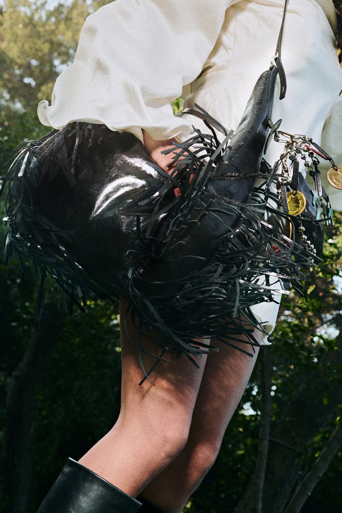 A person in a white outfit holds a glossy black fringed INA KENT handbag with gold charms, standing outdoors with greenery in the background. Black boots are partially visible.