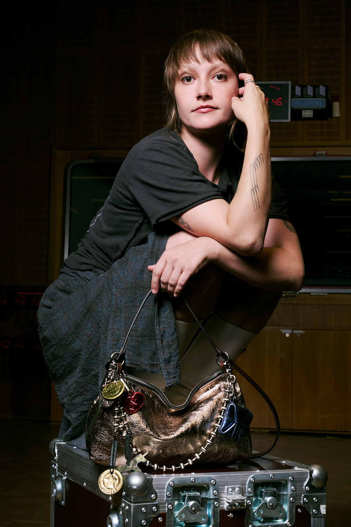 Person with short hair crouches on a metal trunk, resting an arm on their knee, with an INA KENT metallic handbag adorned with keychains in front, in a room with wood panel walls and studio equipment.