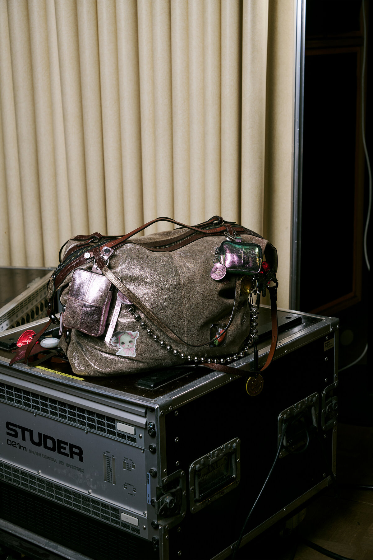 A worn brown INA KENT leather bag with keychains sits on a black Studer equipment case in front of beige curtains.