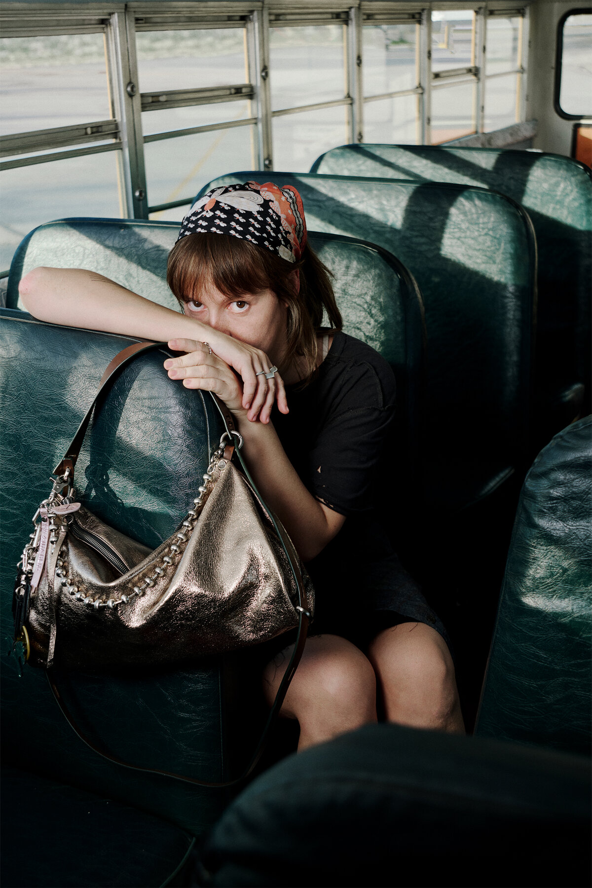 A person with a patterned headscarf sits on a green school bus seat, resting their arm on the seat back and holding a large metallic INA KENT handbag.