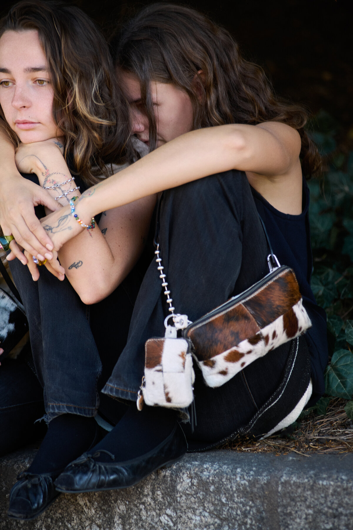 Two people sit close together on a curb, both dressed in black. One rests their head on the other’s shoulder, carrying a small cowhide-patterned INA KENT bag.