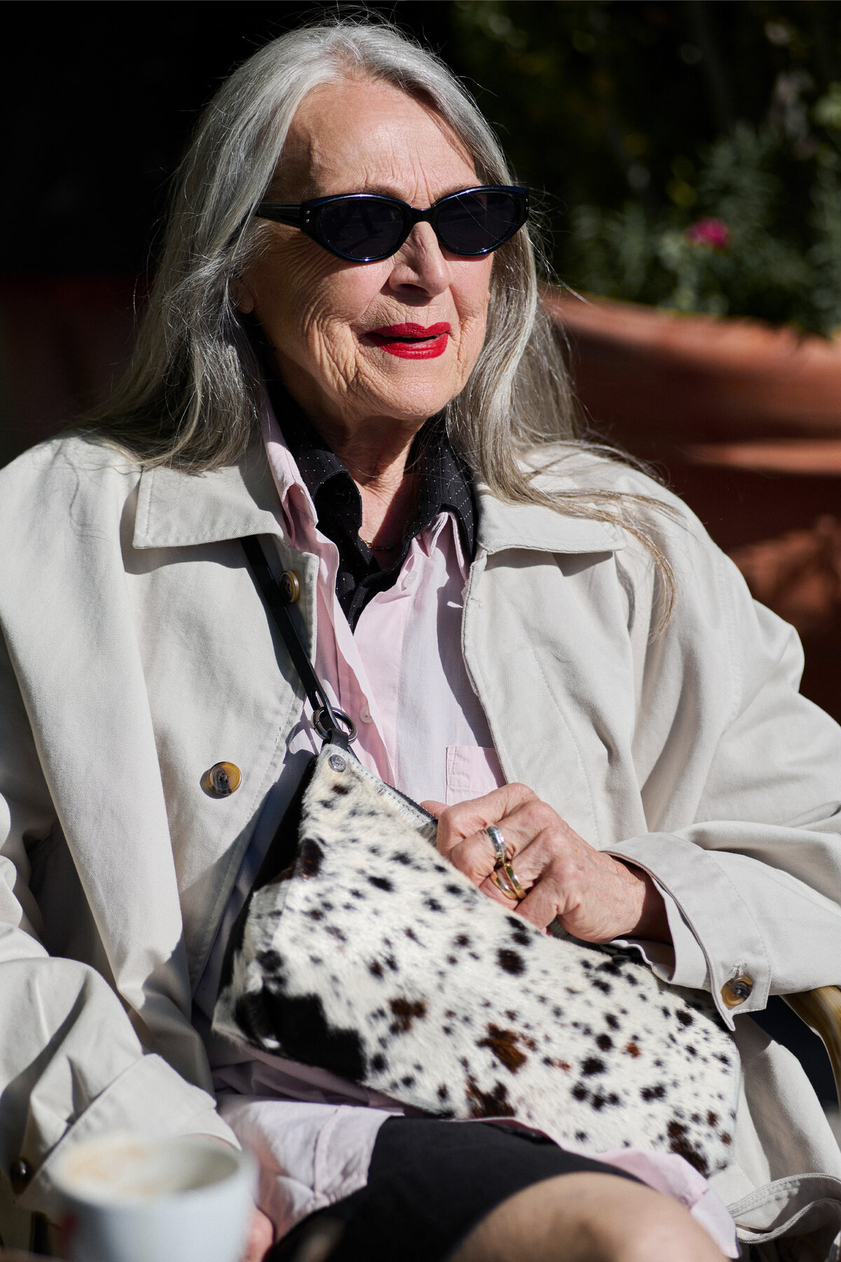 An older woman with long gray hair, wearing sunglasses and a light-colored trench coat, holding a spotted print clutch bag.