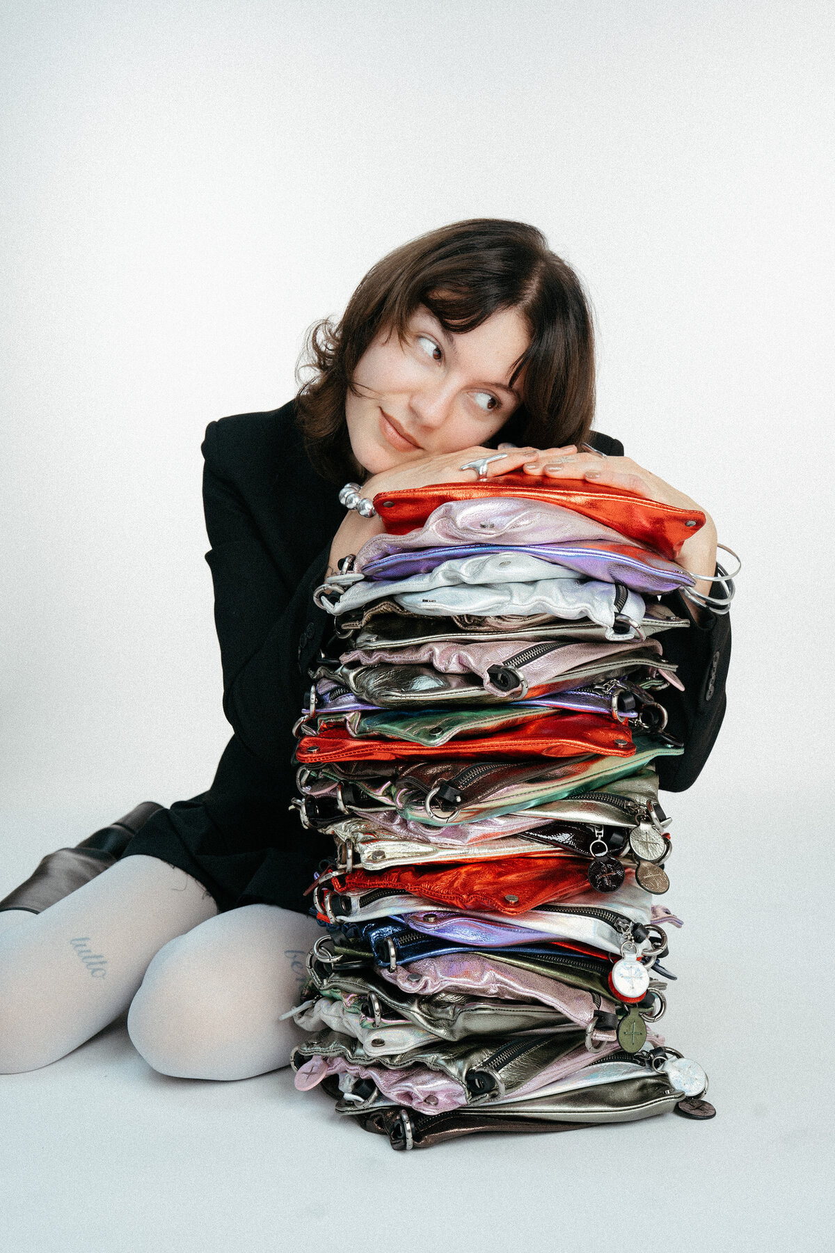 A person in a black blazer rests their chin on a tall stack of colorful, metallic zippered pouches.