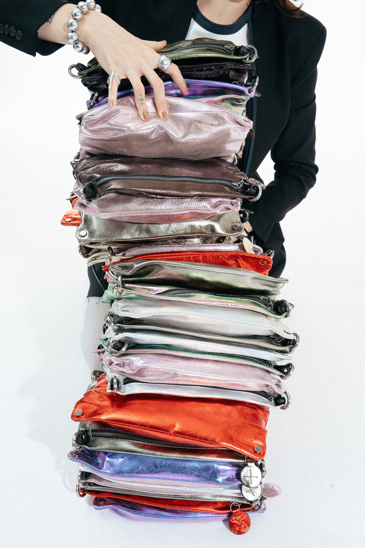 A person in a black blazer holds a tall, precarious stack of metallic leather crossbody bags.