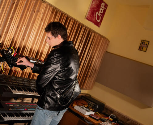 Eine Person steht in einem Musikstudio, trägt eine schwarze Lederjacke von INA KENT und bedient ein Synthesizer-Keyboard, umgeben von Audiogeräten vor dem Hintergrund einer hölzernen Akustikplatte.