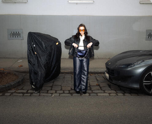 Eine Person mit Sonnenbrille und schwarzem Outfit steht auf einem gepflasterten Gehweg zwischen einem überdachten Objekt und einem geparkten Auto und hält eine geöffnete INA KENT-Tasche in der Hand.
