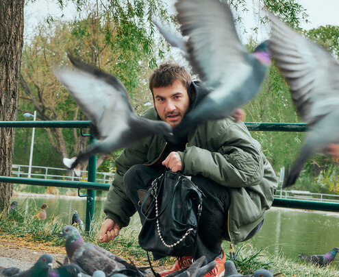 Ein Mann hockt in der Nähe eines Teiches, umgeben von Tauben, wobei mehrere Vögel im Flug sind - einer hält eine INA KENT-Tasche fest - und sein Gesicht teilweise verdecken.