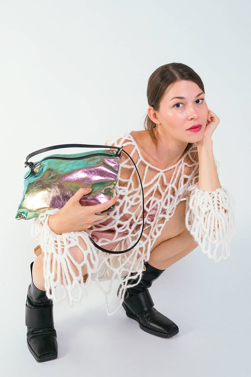 A woman in a white, netted outfit and black boots crouches, holding a shiny INA KENT bag with multicolored accents, looking directly at the camera.