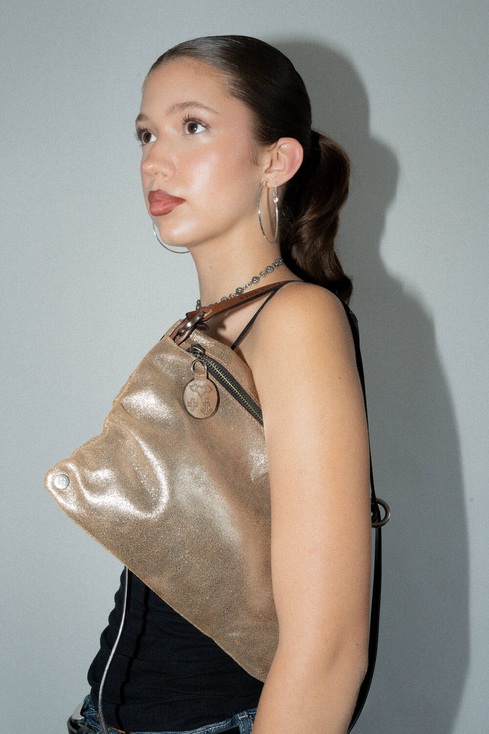 A person with hoop earrings and a sleek ponytail poses against a gray background, wearing a black top and an INA KENT metallic gold sling bag.