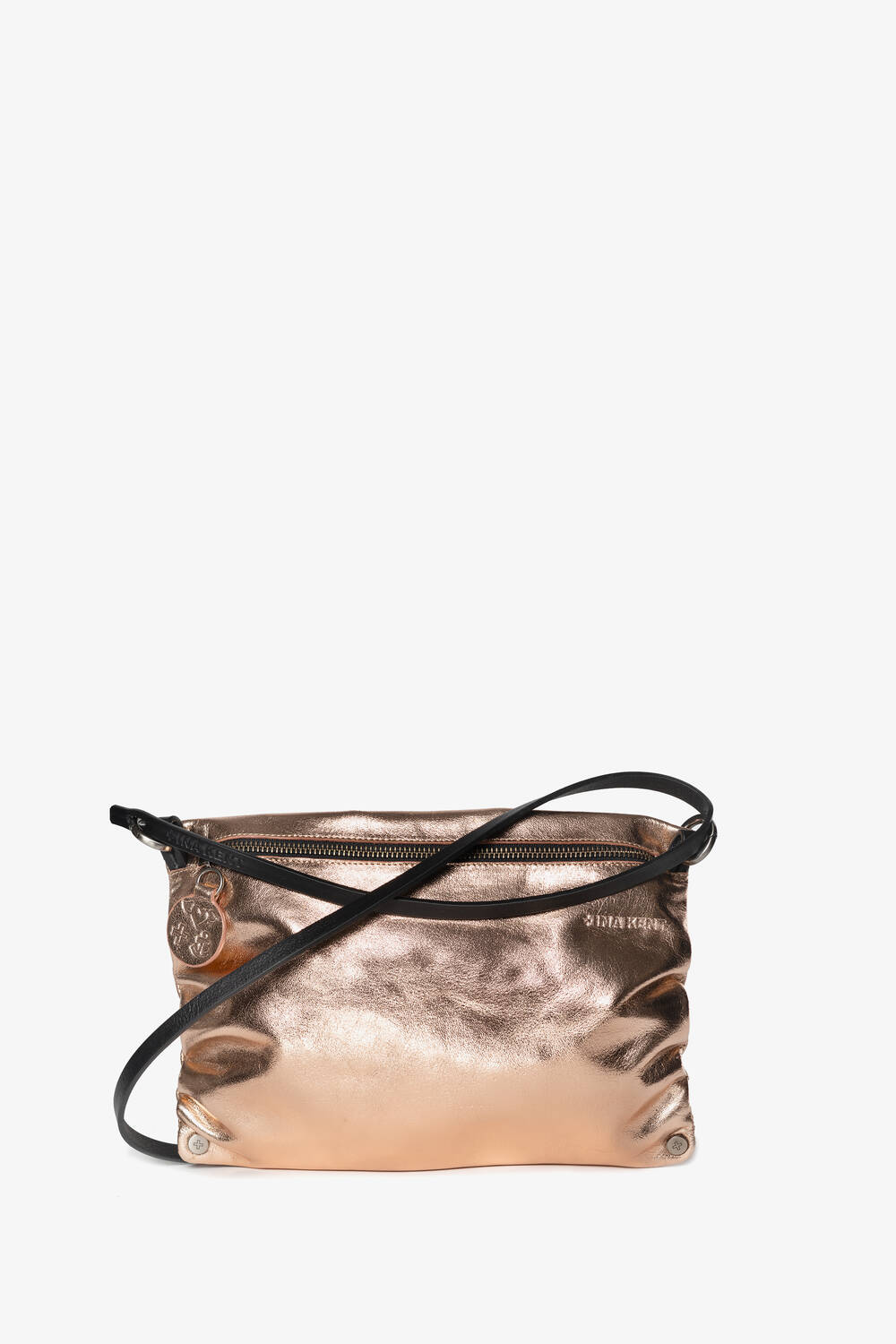 A small metallic rose gold crossbody bag by INA KENT with a black strap and zipper closure. The bag features a slightly crinkled texture and a circular charm hanging from the zipper pull.