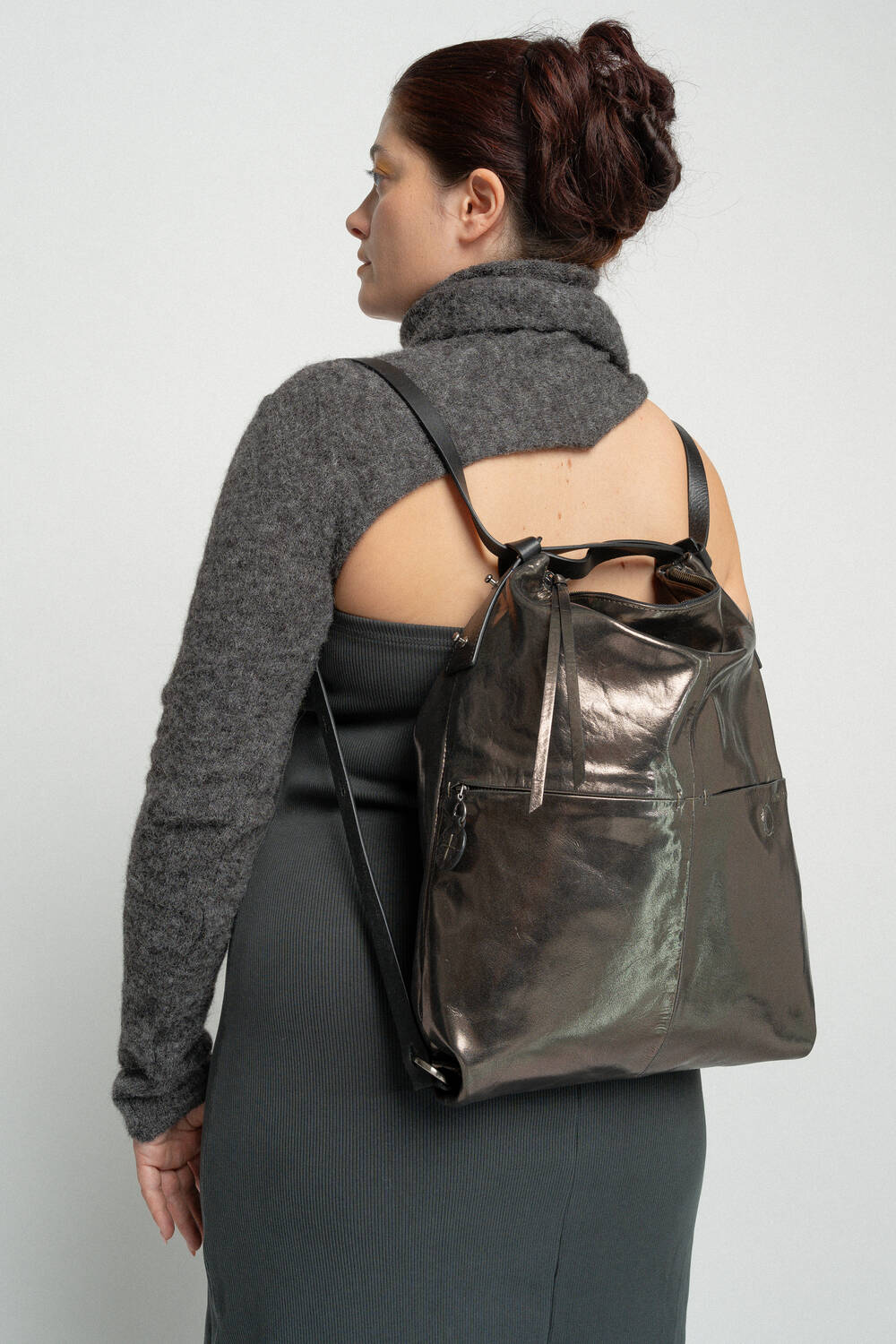 A person with dark hair in a bun, wearing a gray sweater and dark dress, is seen from behind, showcasing an INA KENT metallic gray backpack.