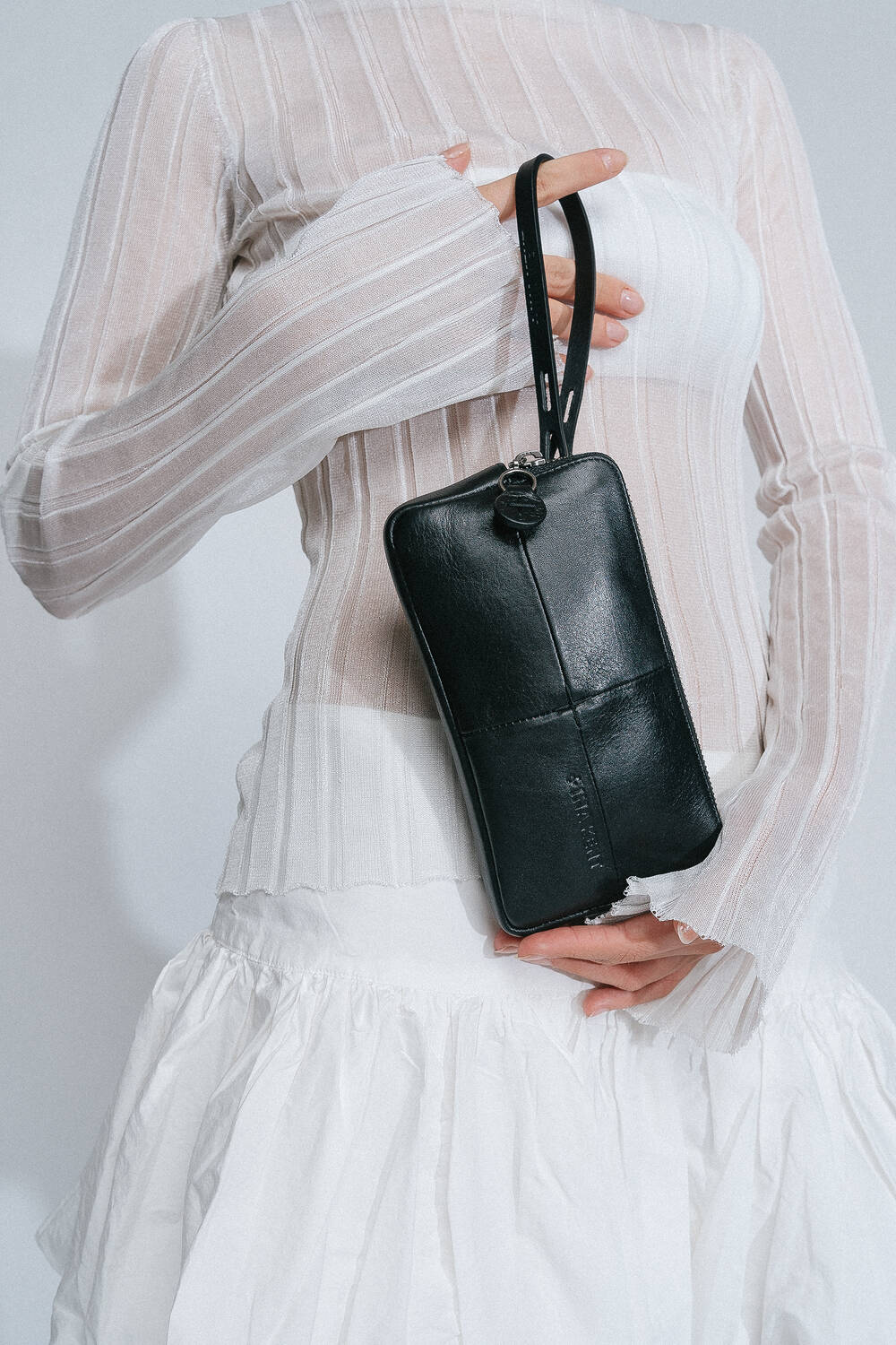 A person in a sheer, long-sleeve top and white skirt holds an INA KENT black wristlet purse.