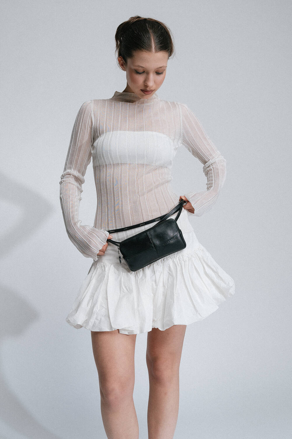 A woman wearing a sheer long-sleeved top and a white skirt looks down at her INA KENT black belt bag around her waist.