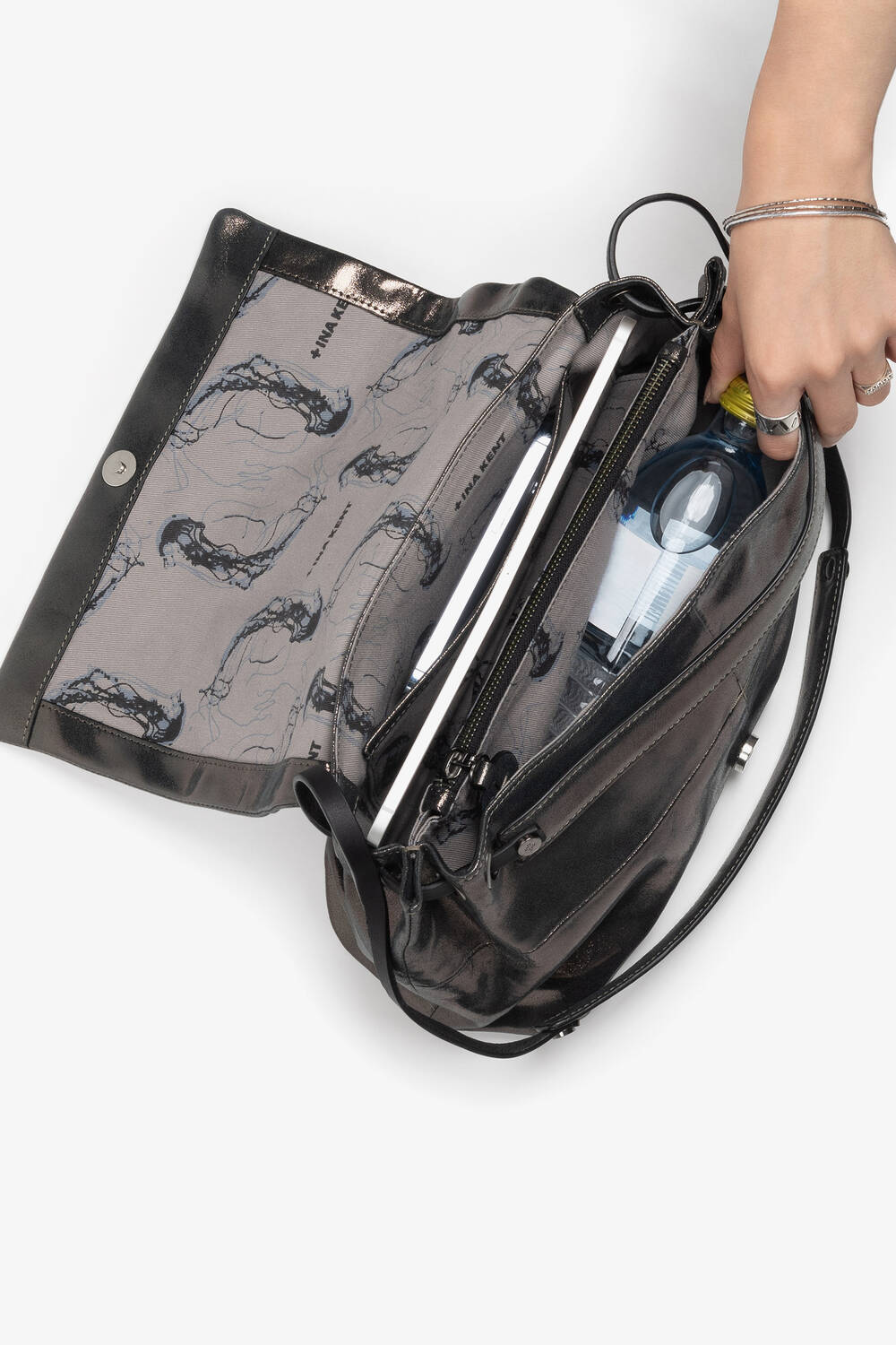 A person is opening a black INA KENT handbag, revealing its contents which include a water bottle, a hairbrush, and an inner zip pocket with a metallic finish.