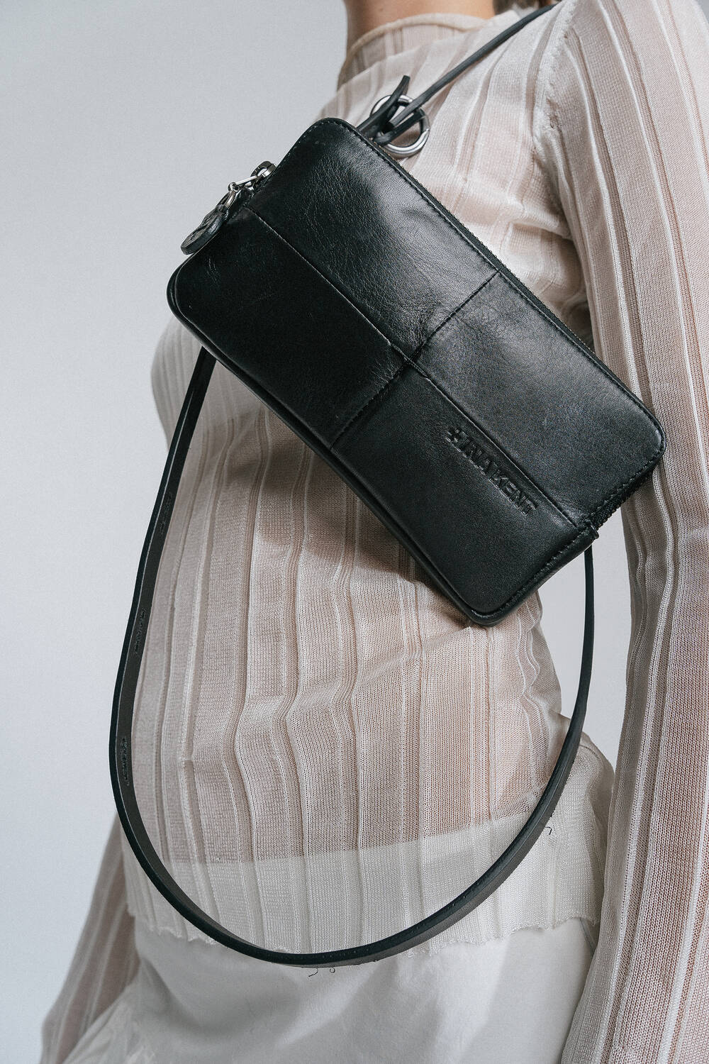 A person is wearing a long sheer white top with an INA KENT black leather crossbody bag slung over their shoulder.