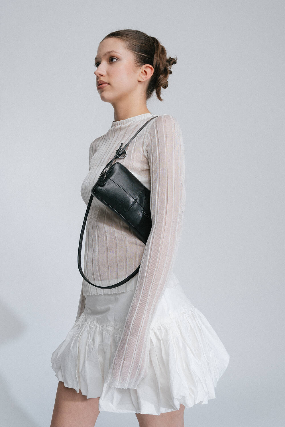 A woman in a sheer long-sleeve top and white skirt poses with an INA KENT black crossbody bag against a plain backdrop.