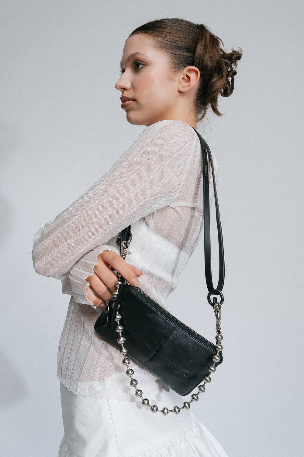 A person with hair tied up, wearing a white top and skirt, holds an INA KENT black handbag over their shoulder.