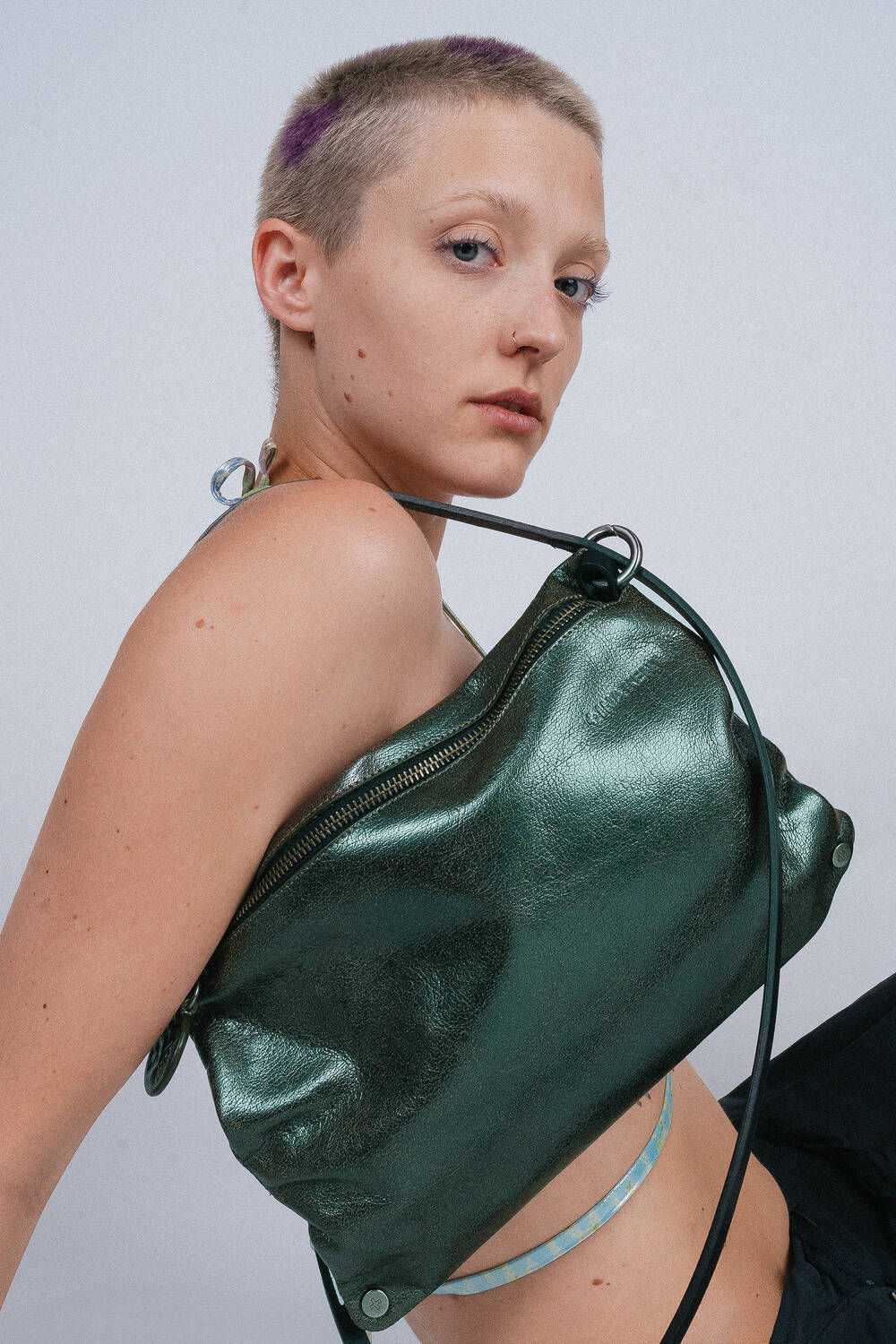 A person with short hair is seen gazing at the camera, their style accentuated by an INA KENT metallic green bag draped over one shoulder.