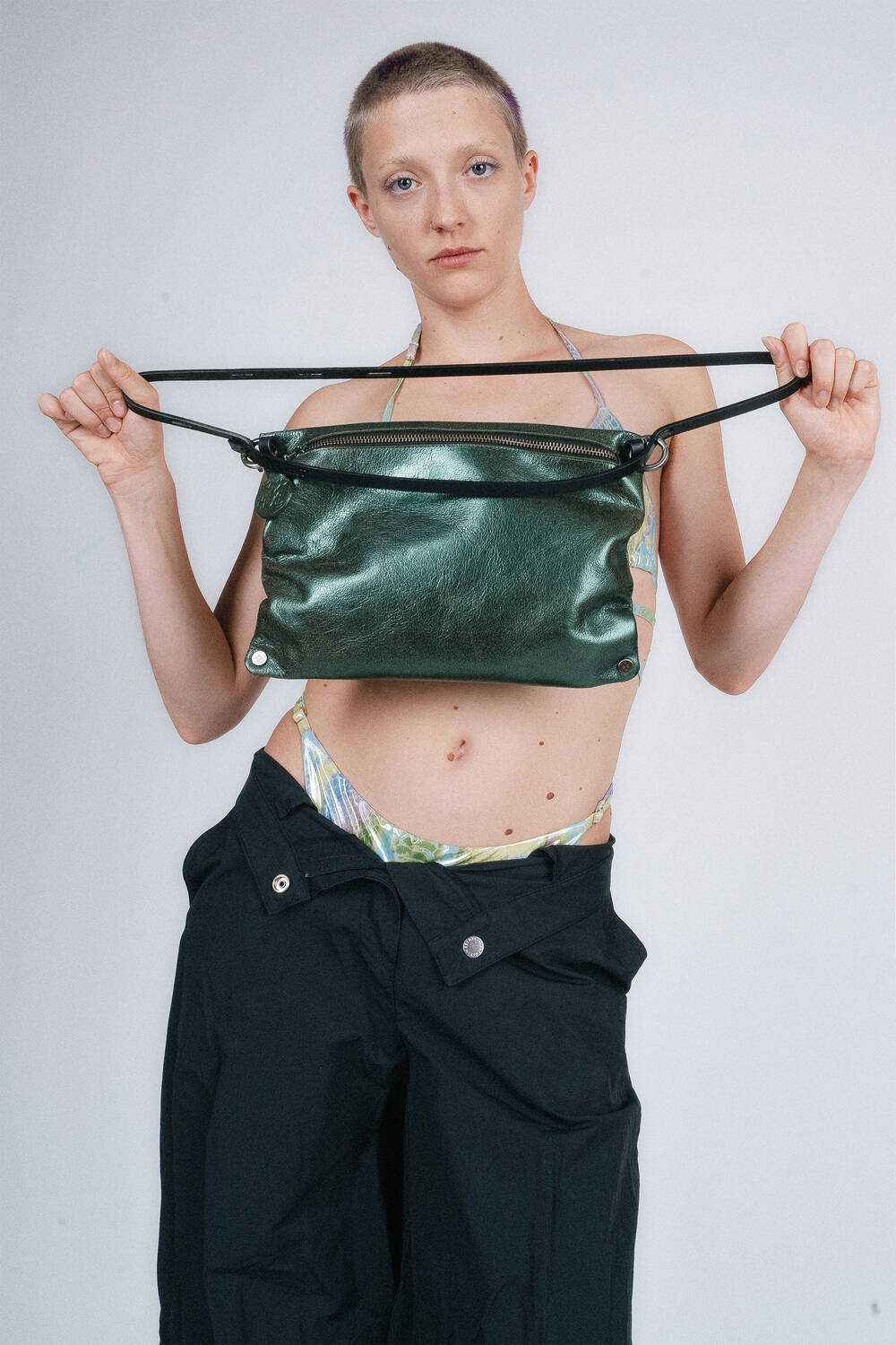 Person holding an INA KENT green leather bag by its straps, wearing dark pants and a colorful top, against a plain background.