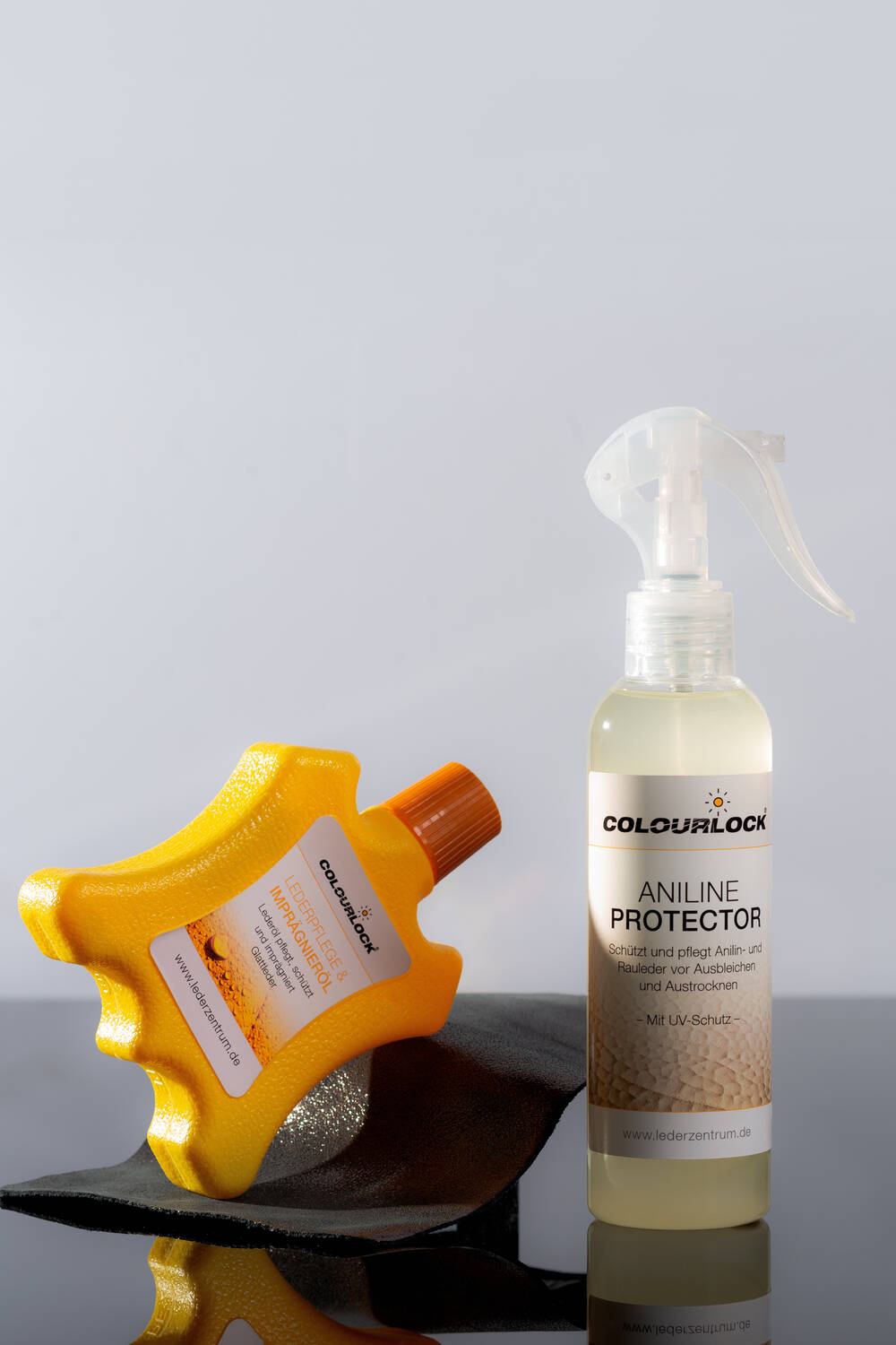 Auf einer reflektierenden Oberfläche stehen zwei Flaschen mit Lederpflegeprodukten; eine ist ein gelber Reinigungsschaum und die andere, mit der typischen Handschrift von INA KENT, ein Spray mit der Aufschrift „Aniline Protector“ von Colourlock.