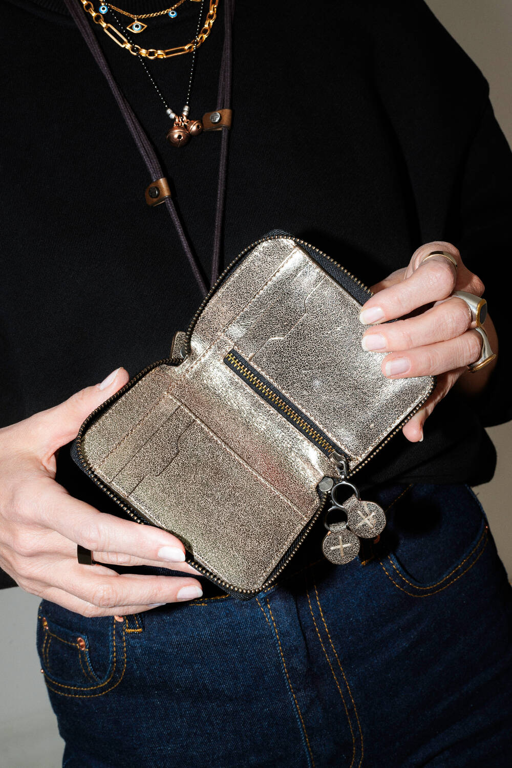 Person holding an open, empty INA KENT metallic wallet, wearing denim pants and layered necklaces.