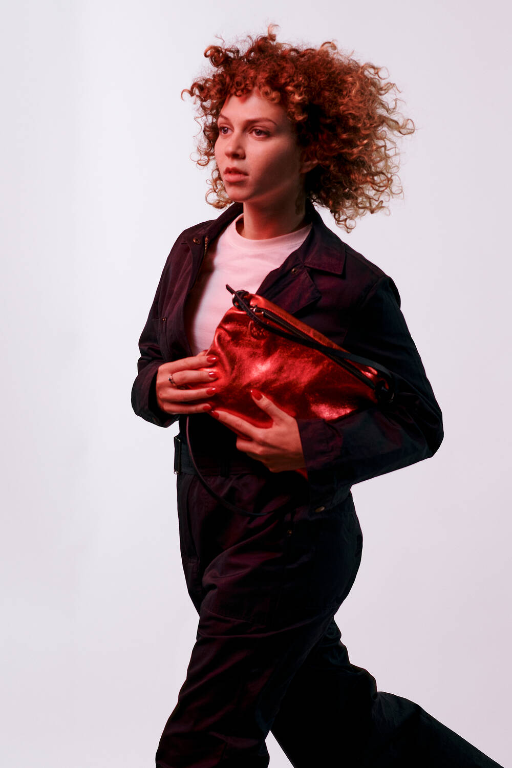 A person with curly hair in a dark outfit strikes a pose against a plain background, showcasing a striking red INA KENT bag.
