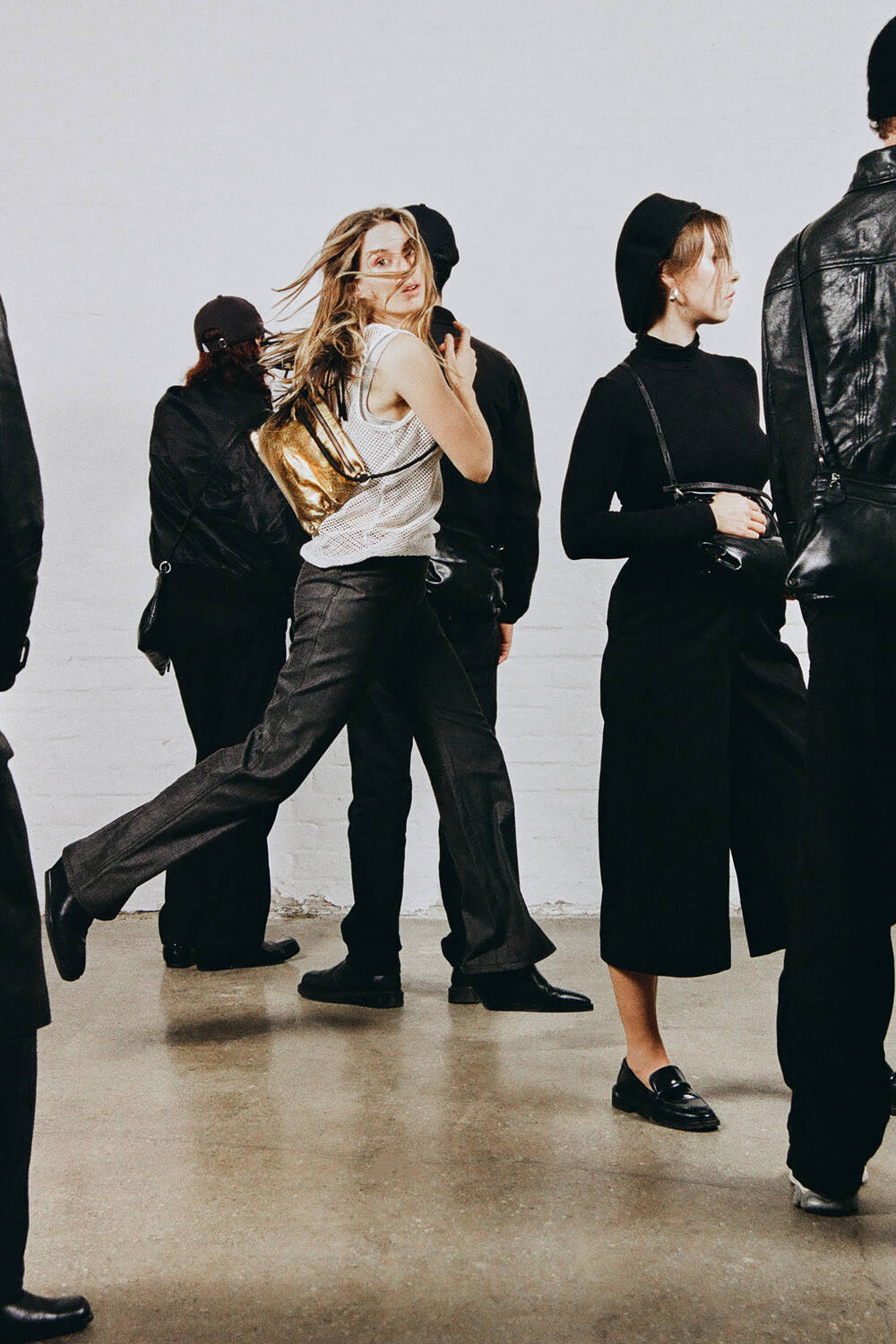 A woman in a white tank top and gray pants walks energetically with her INA KENT gold backpack, surrounded by people dressed in black against a plain white wall.