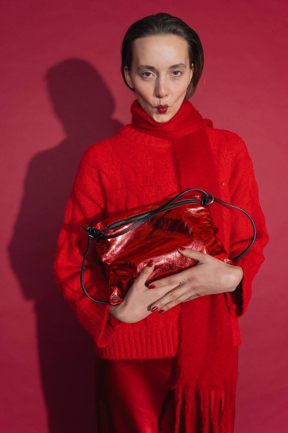 Woman in a red outfit with sweater, scarf and skirt poses in front of a red background. She holds the INA KENT MOONLIT ed.1 bag in shiny metallic chili-red leather in front of her upper body.