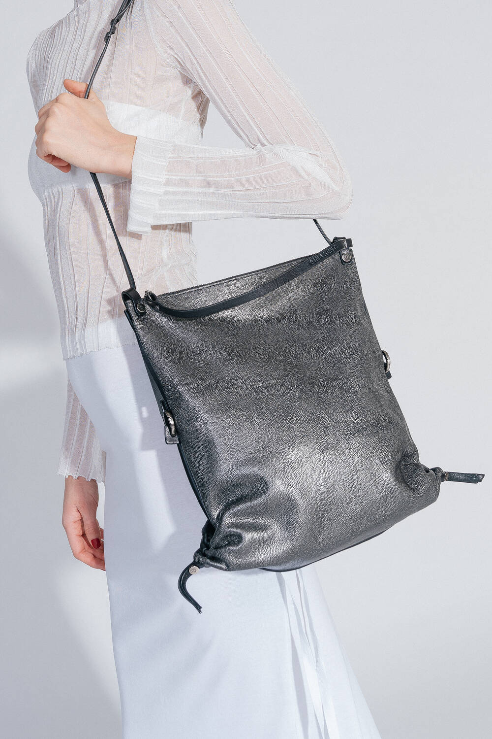 Frau in einem weißen, durchscheinenden Oberteil und weißem Rock trägt die INA KENT-Tasche AD LIB ed.1 über der Schulter. Die Tasche hat eine strukturierte, schwarze Oberfläche und wird als Tote Bag am liegen Riemen getragen.