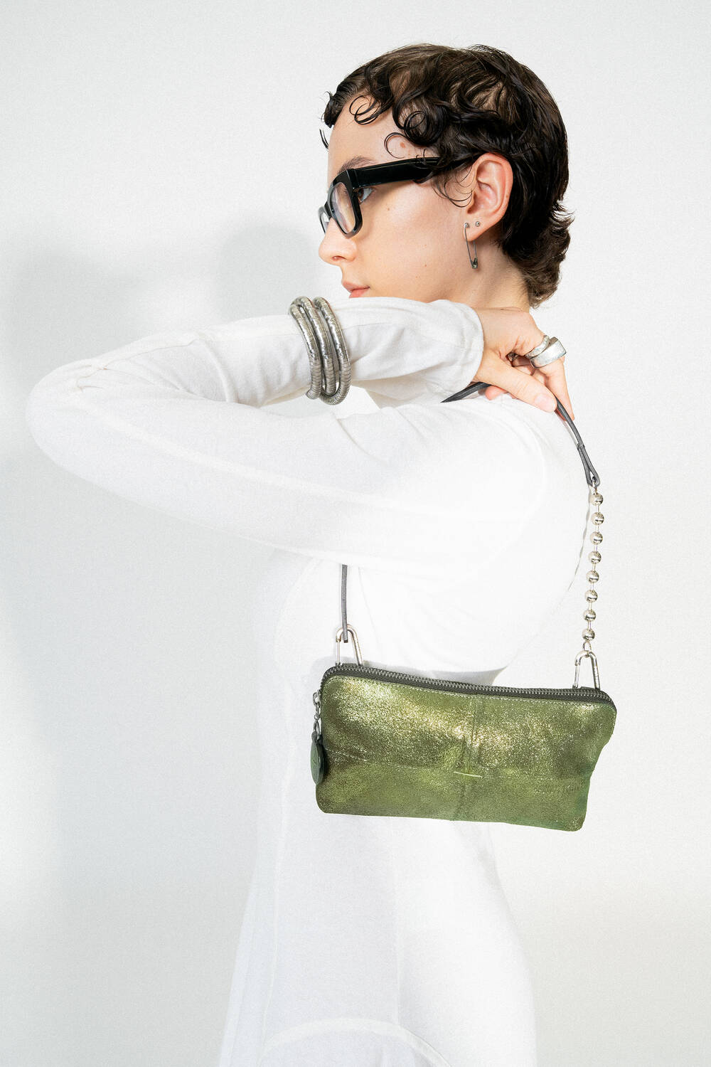 Seitenansicht einer weiblich gelesenen Person, die die INA KENT Tasche IVI ed.3 in crackled guacamole Leder über der Schulter trägt. Die Tasche hat eine silberne Kugelkette und wirkt auffällig durch ihren grünen Metallic-Glanz.