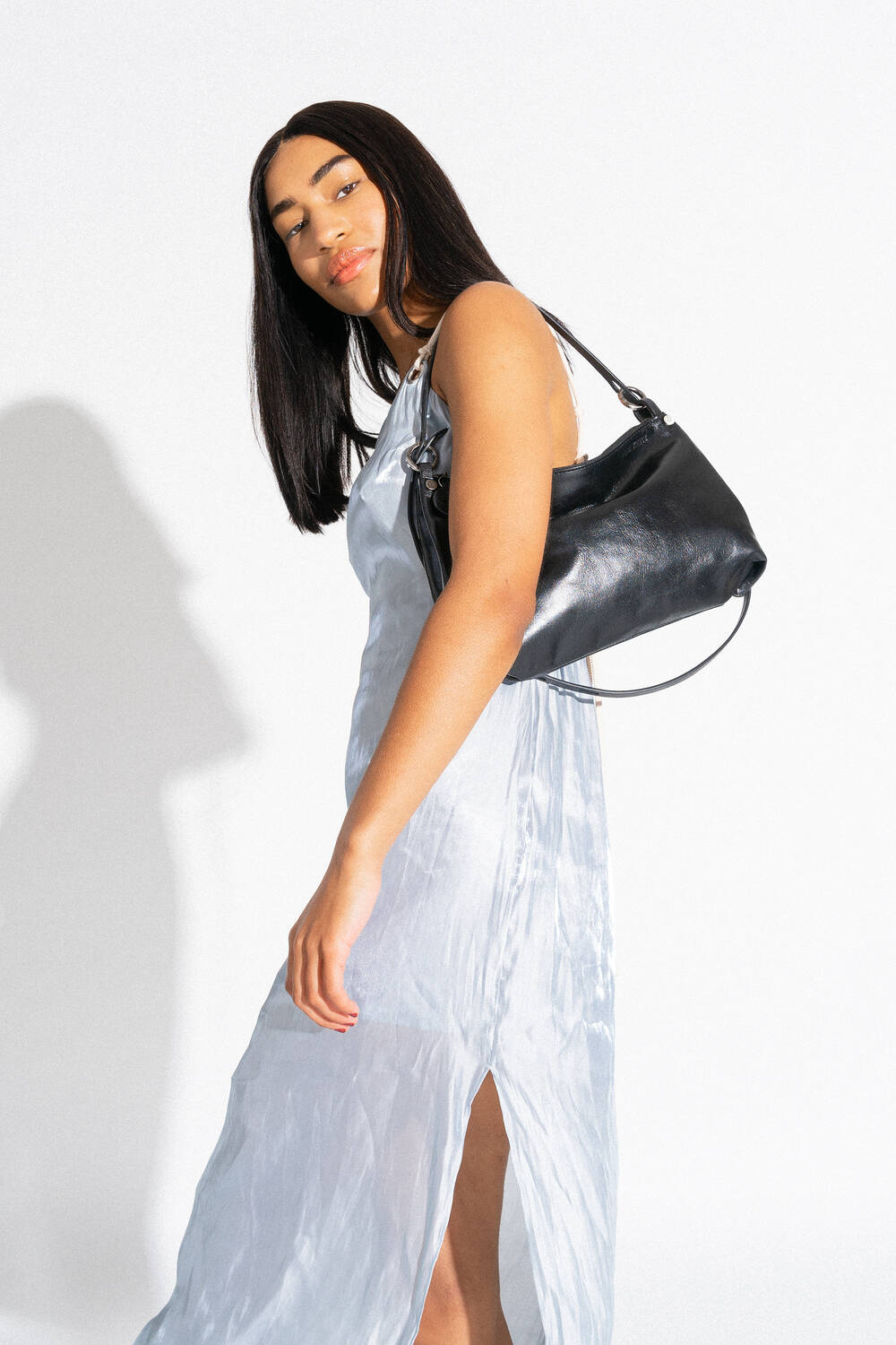 Eine Frau in einem hellblauen Kleid trägt eine schwarze Handtasche von INA KENT auf ihrer Schulter und steht vor einem schlichten weißen Hintergrund.