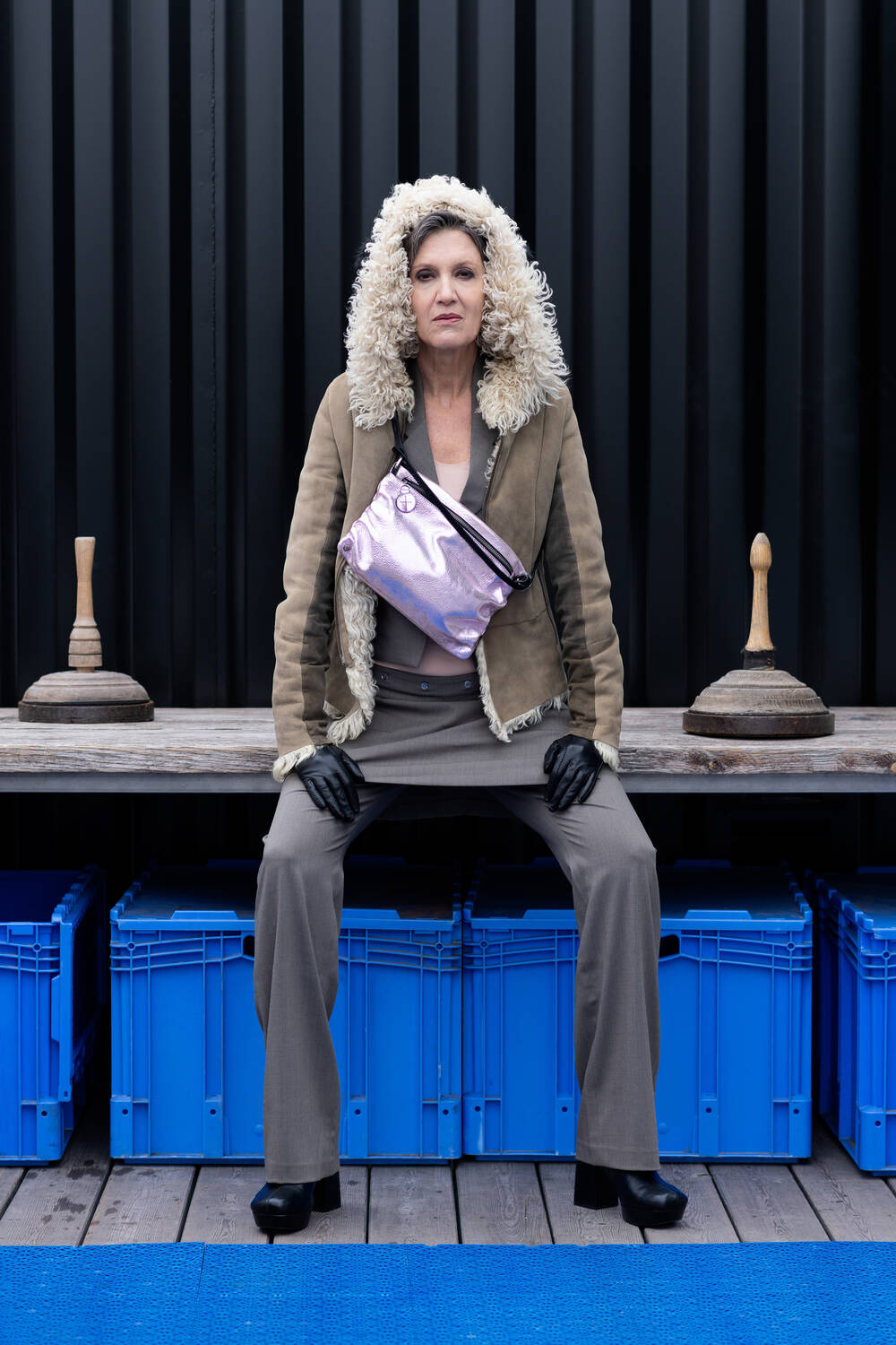 Die Frau sitzt auf einer Holzbank vor einer schwarzen Containerwand mit blauen Kisten darunter. Sie trägt eine metallic-lavender-rose Hüfttasche von INA KENT diagonal.