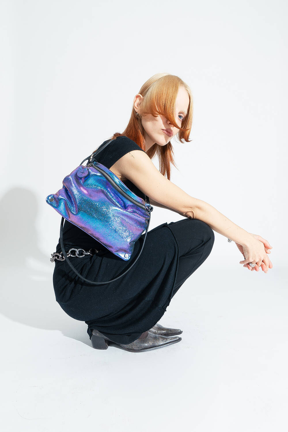 Eine Person in schwarzer Kleidung posiert vor einem weißen Hintergrund mit der zweifarbigen metallic Leder-Crossbody-Tasche MOONLIT ed.1 in Lila-blau 